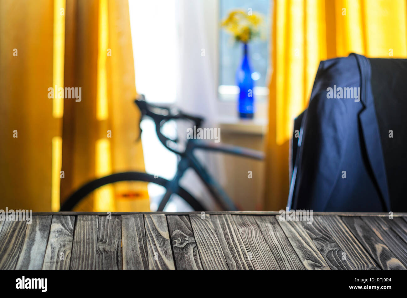 Product Placement Background. Wooden Table Against Blurry Interior of Cyclist's Living Room with Large Window and Blue Sport Bicycle. Blue Jacket on t Stock Photo