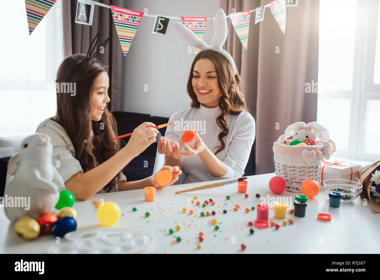 Mother and daughter prepare for Easter. Young woman wear rabitt ears and hold egg. Girl painting it. They have fun together. Daylight Stock Photo