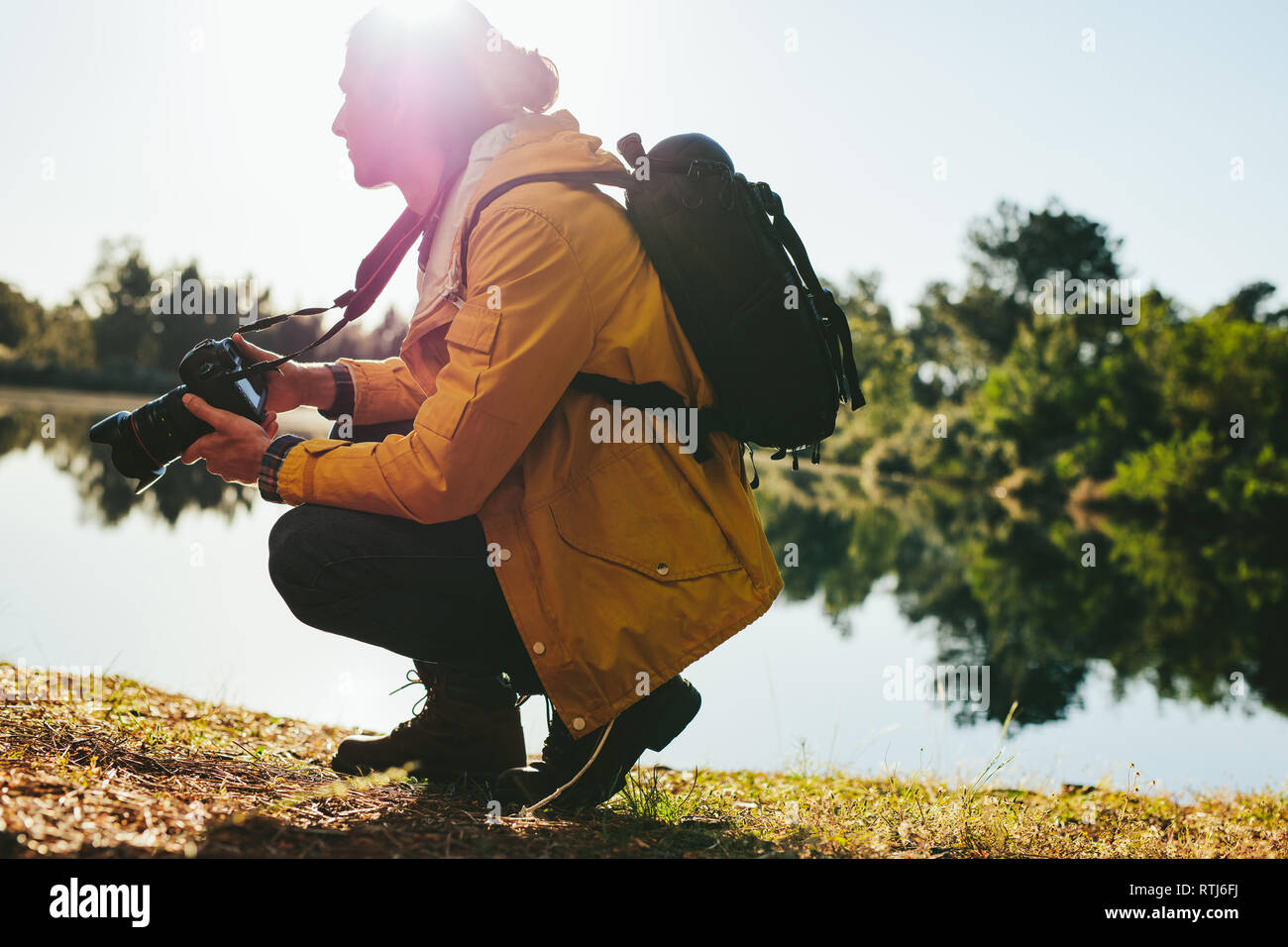 Side view of a traveler sitting beside a lake holding a digital camera. Man shooting photos of nature sitting beside a lake on a sunny day. Stock Photo