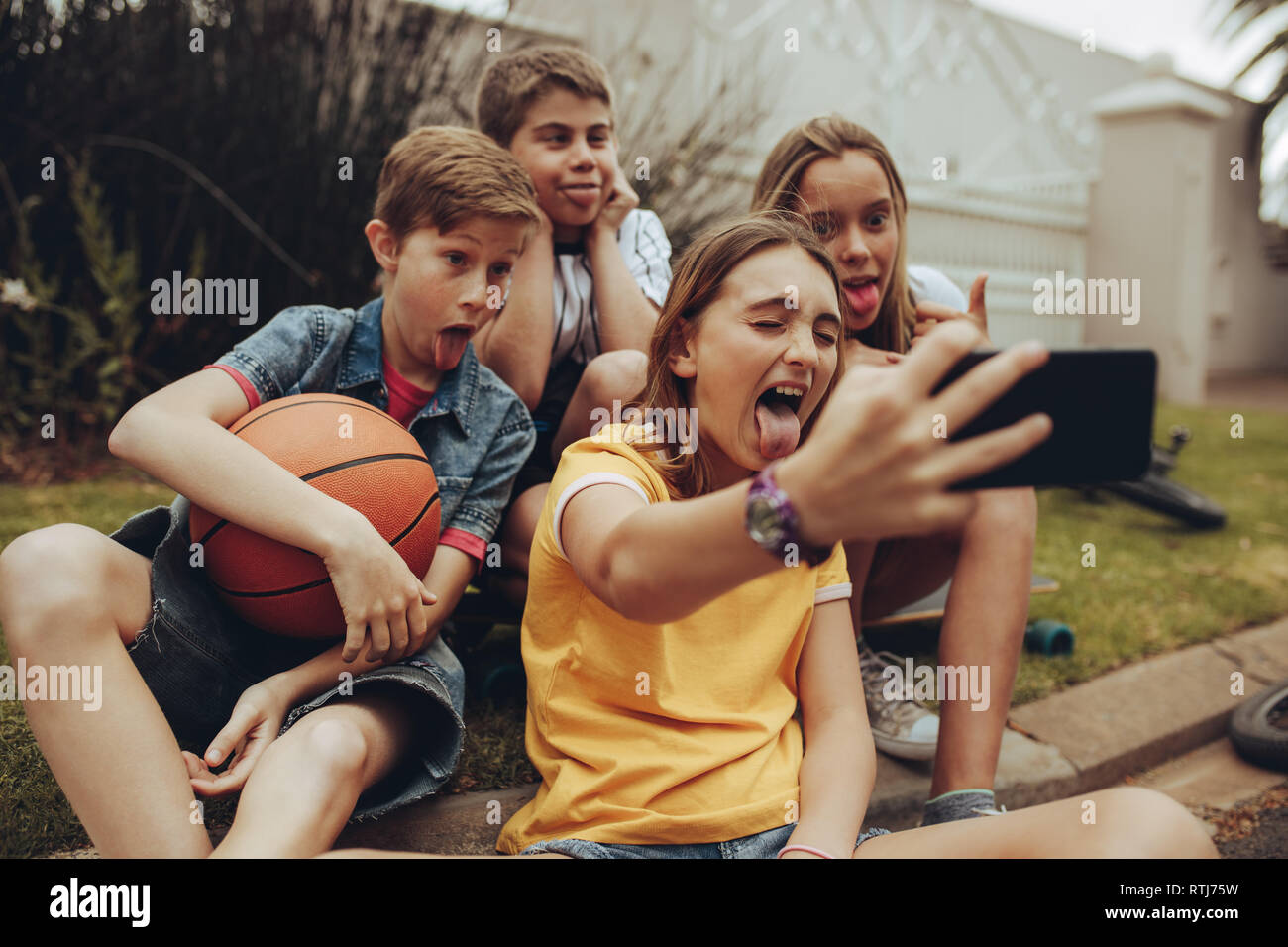 Kids making funny faces and sticking out their tongue for a selfie. Girl taking a selfie with her friends sitting outdoors. Stock Photo