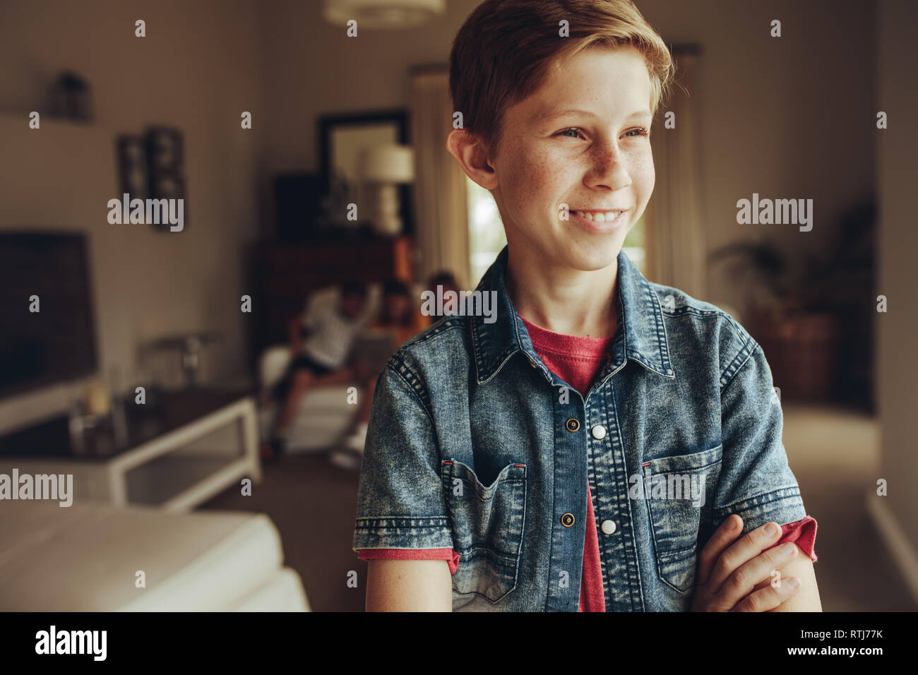 Portrait of a smiling boy standing at home with arms crossed. Boy standing at home and looking away while his friends sitting together in the backgrou Stock Photo