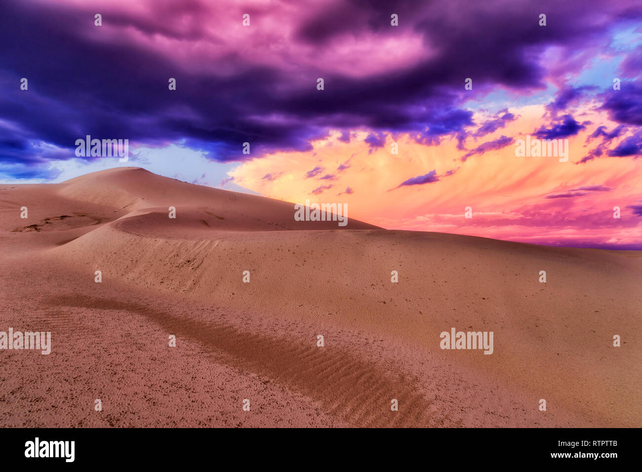 Vivid colourful stormy clouds over arid sandy desert with ground surface punched by drops of rain at sunset. Stock Photo