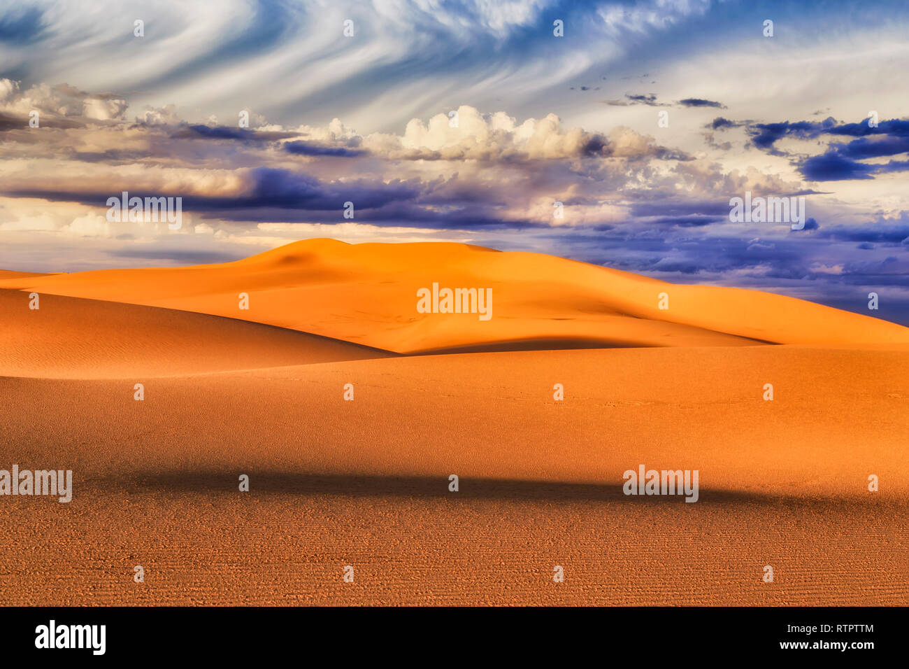 Endless lifeless sand dunes under clouds and blue sky in soft sunset light with shades drawing images with shapes of sand masses. Stock Photo