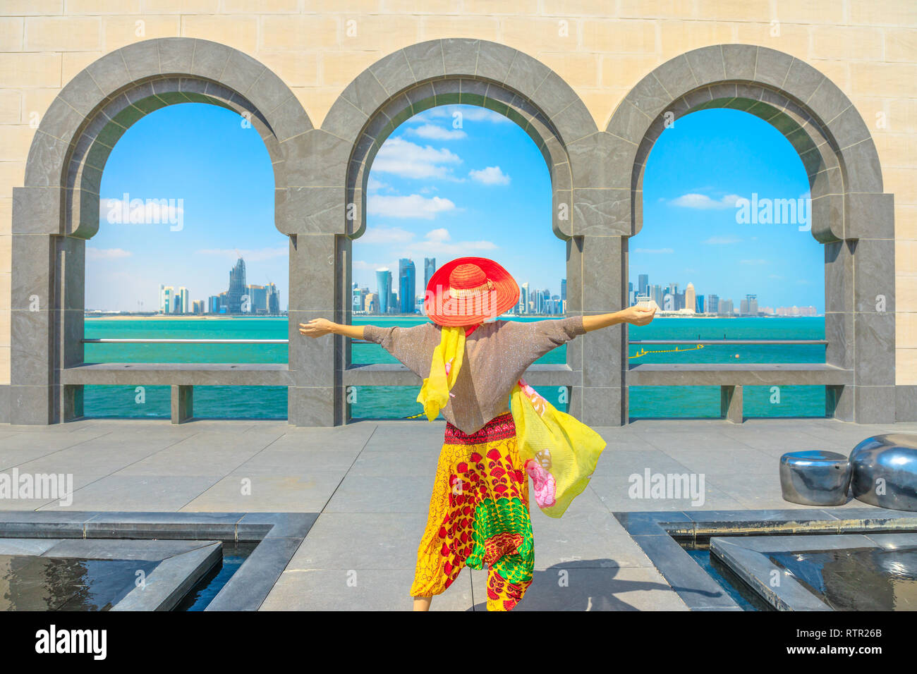 Carefree caucasian woman with sunhat looking skyscrapers of Doha West Bay skyline through arches of famous place. Traveler tourist enjoys in Qatar Stock Photo