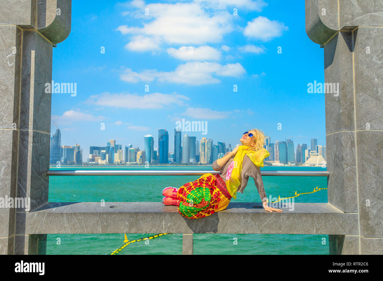 Happy blonde woman sitting under arches enjoying the view of Doha West Bay skyline outdoors of Museum of Islamic Art. Caucasian tourist in Qatar Stock Photo