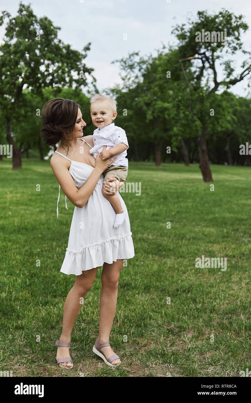 Beautiful and happy young mom in short white dress with a cute little stylish son on her hands posing at green park Stock Photo Alamy