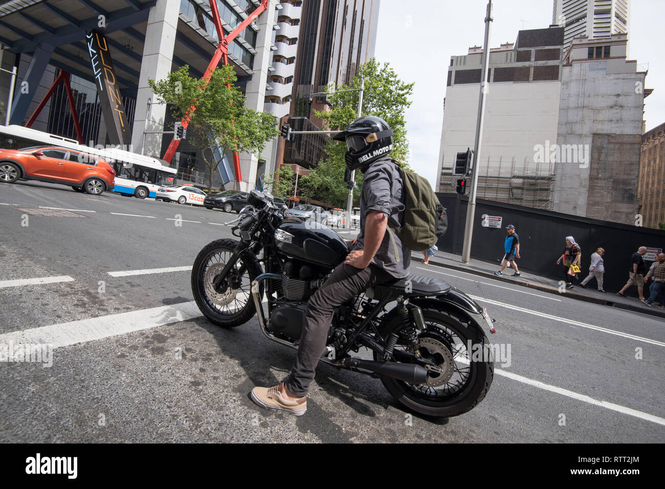 A young motorcyclist in a full face helmet sits and waits at a set of traffic lights in Sydney's CBD in Australia on his restored older motorbike. Stock Photo