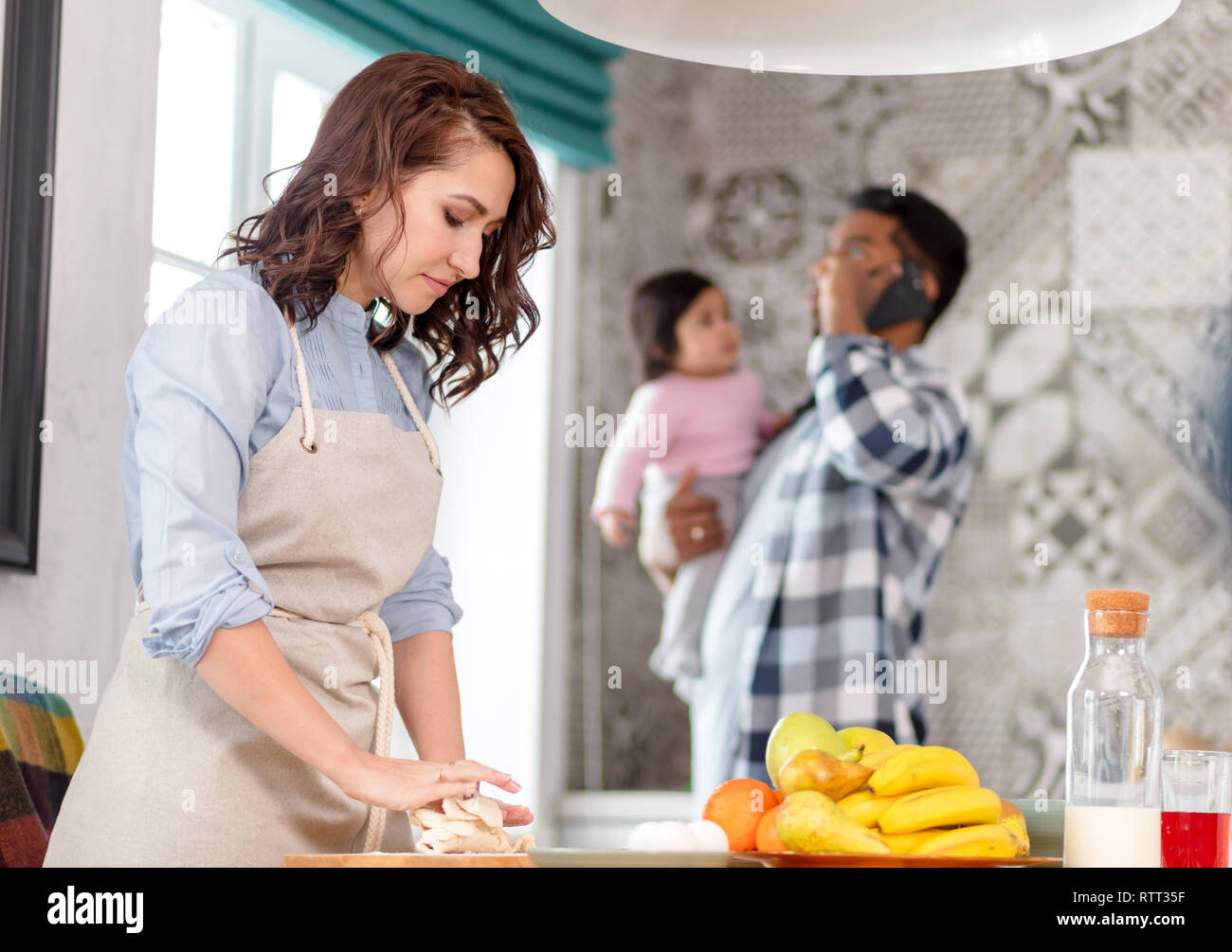 attractive hardworking wife working in the kitchen while her husband holding their child and talking on the phone. close up photo. blurred background. Stock Photo