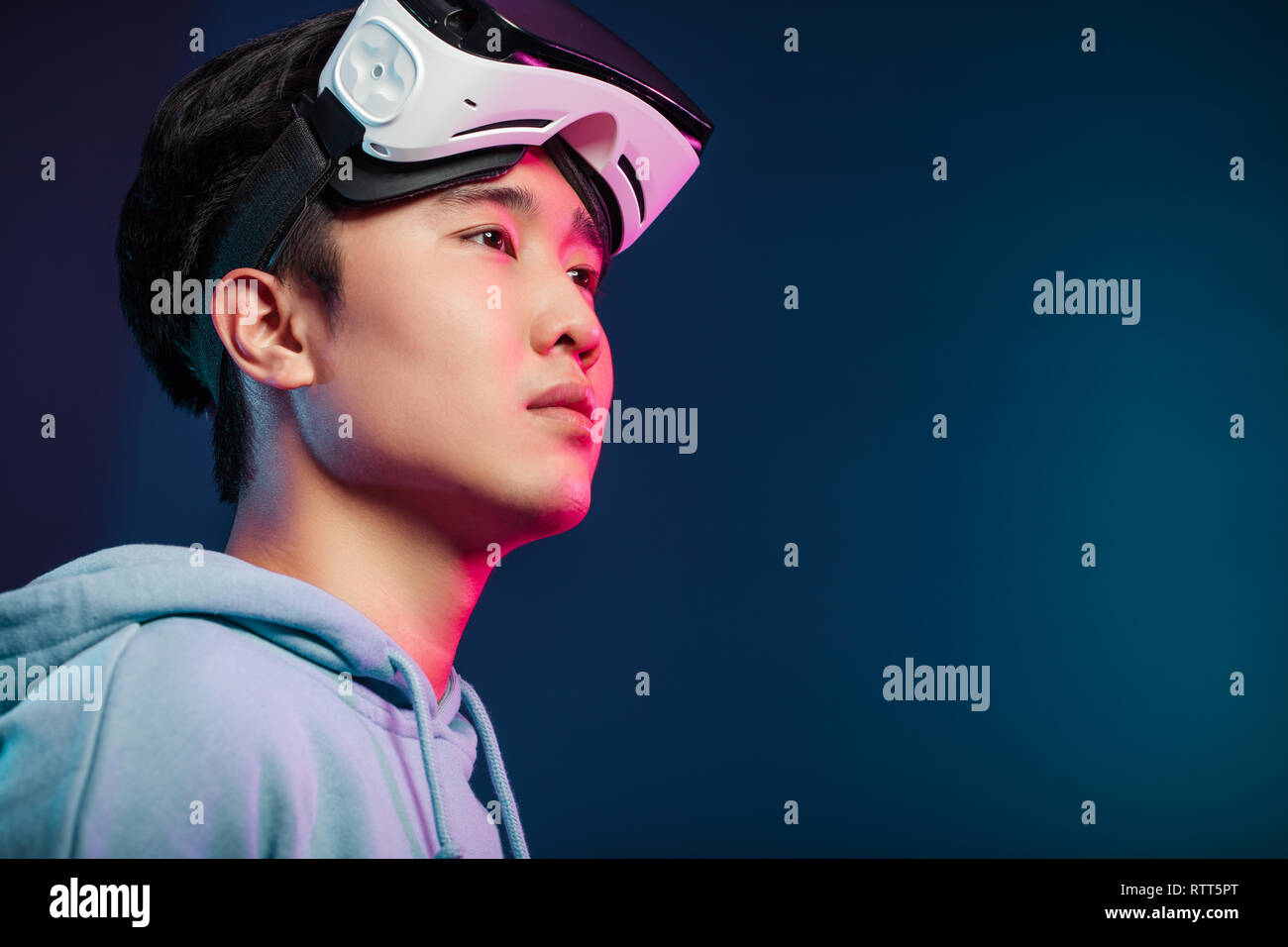 Successful Asian male virtual reality gamer taking off vr headset and looking around at studio, isolated portrait over dark blue background with doubl Stock Photo
