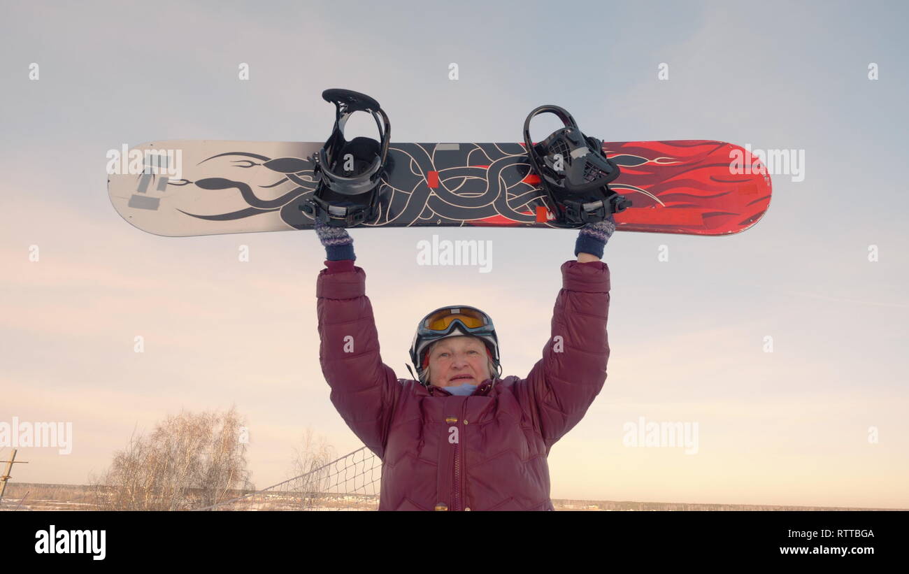 Cheerful older woman rising up snowboard on snowy slope at winter