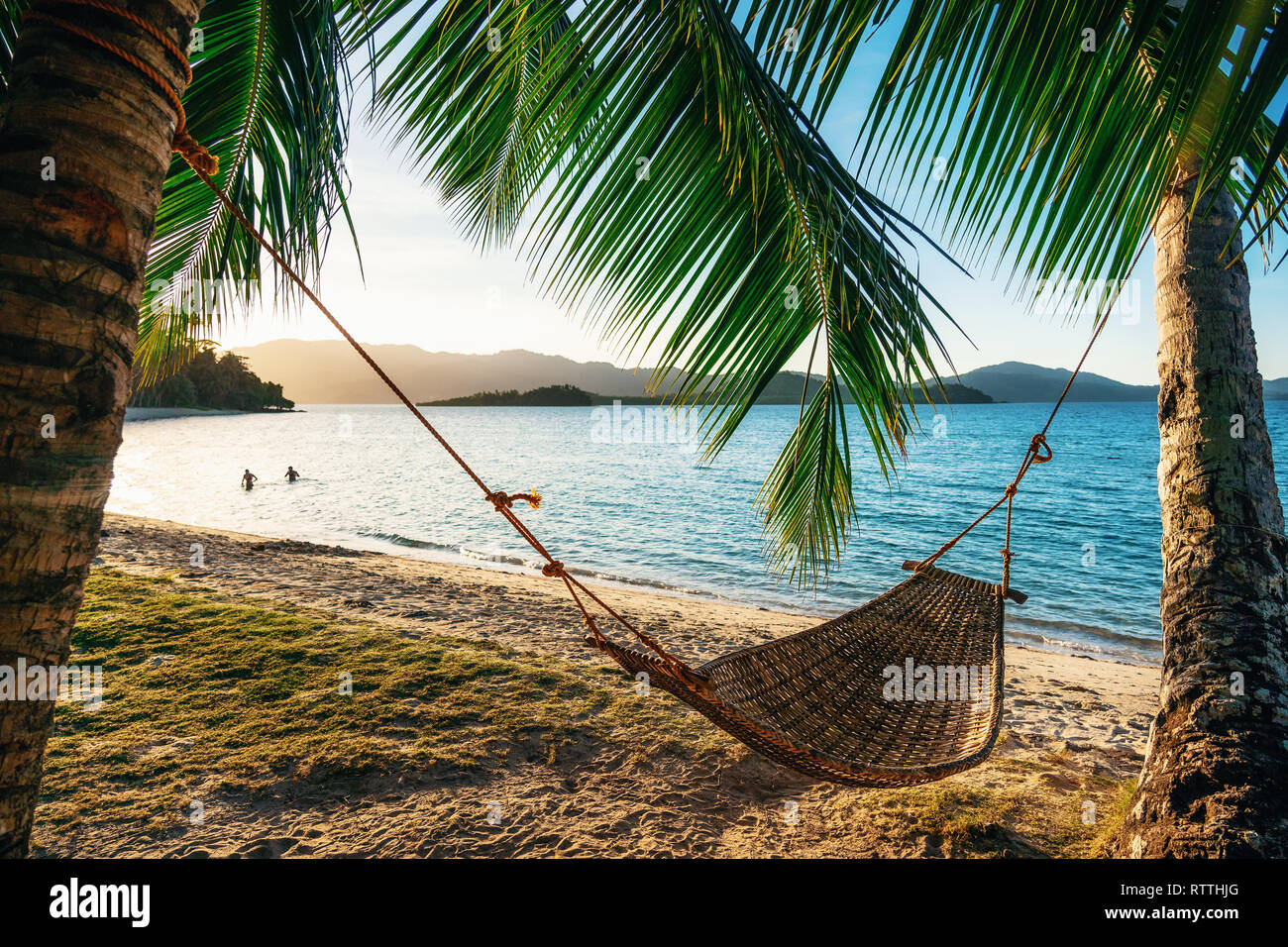 Empty hammock between two palm trees on the beach at sunset. Silhouette of couple in the background in sea. Holiday and vacation concept Stock Photo