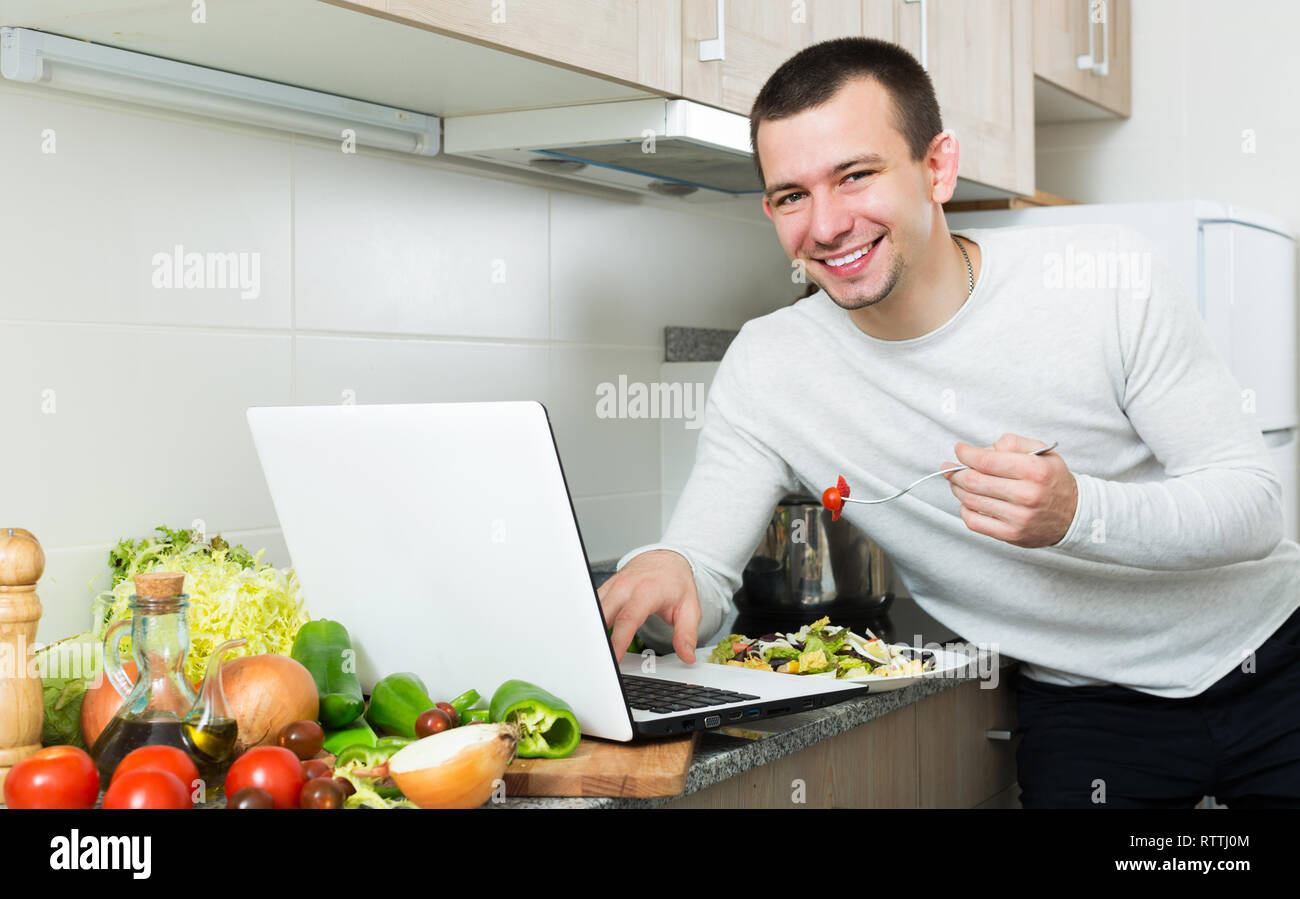 Satisfied male working with laptop holding vegies in the kitchen Stock Photo