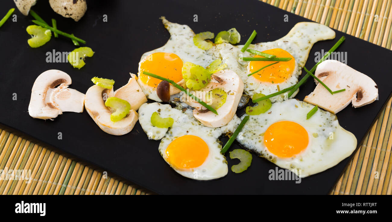 Broken fried quail eggs served on black board with mushrooms, sliced celery and onion Stock Photo