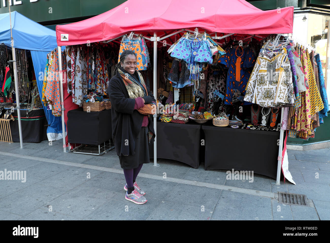 A smiling woman selling African textiles, clothing and jewellery at a street market stall on Brixton Road in Brixton South London UK  KATHY DEWITT Stock Photo