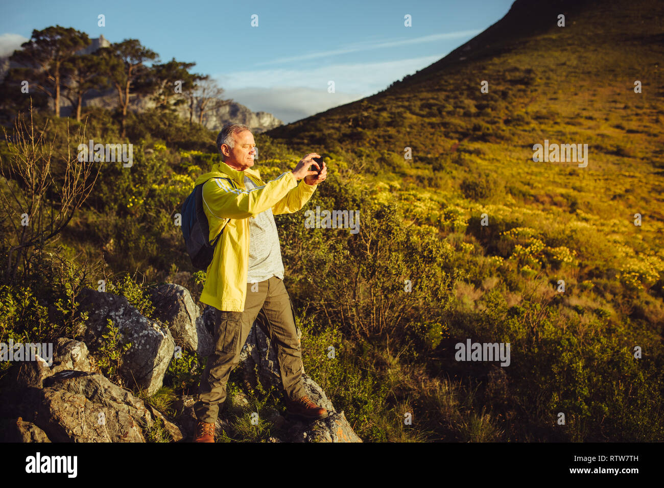 Trekker standing on top of a hill and taking photo using mobile phone. Senior man enjoying trekking adventure capturing beautiful scenes. Stock Photo