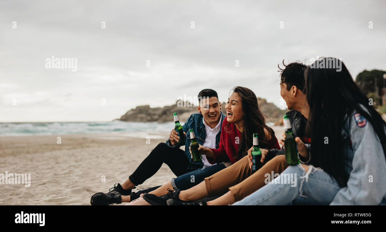 Group of asian men and women sitting at the sea shore with beers. Four young friends partying on the beach. Stock Photo