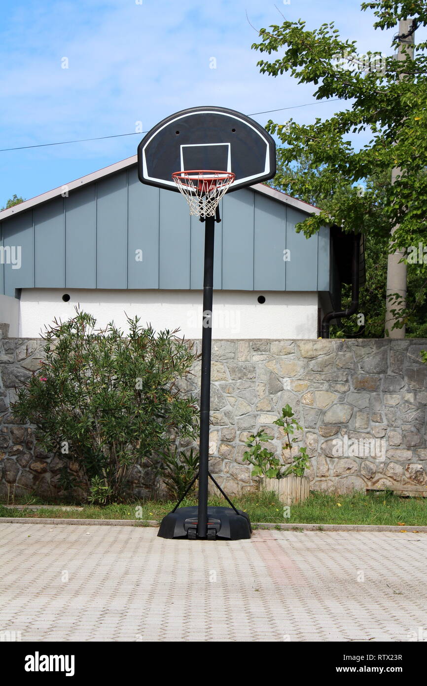Amateur metal and plastic basketball hoop mounted on stone tiles backyard in front of stone wall and outdoor structure surrounded with trees Stock Photo