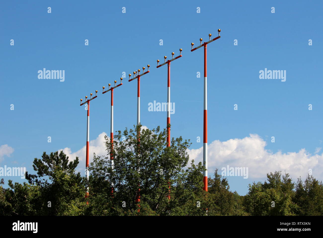 Multiple high metal red and white poles with airport runway guiding lights surrounded with dense trees on cloudy blue sky background Stock Photo