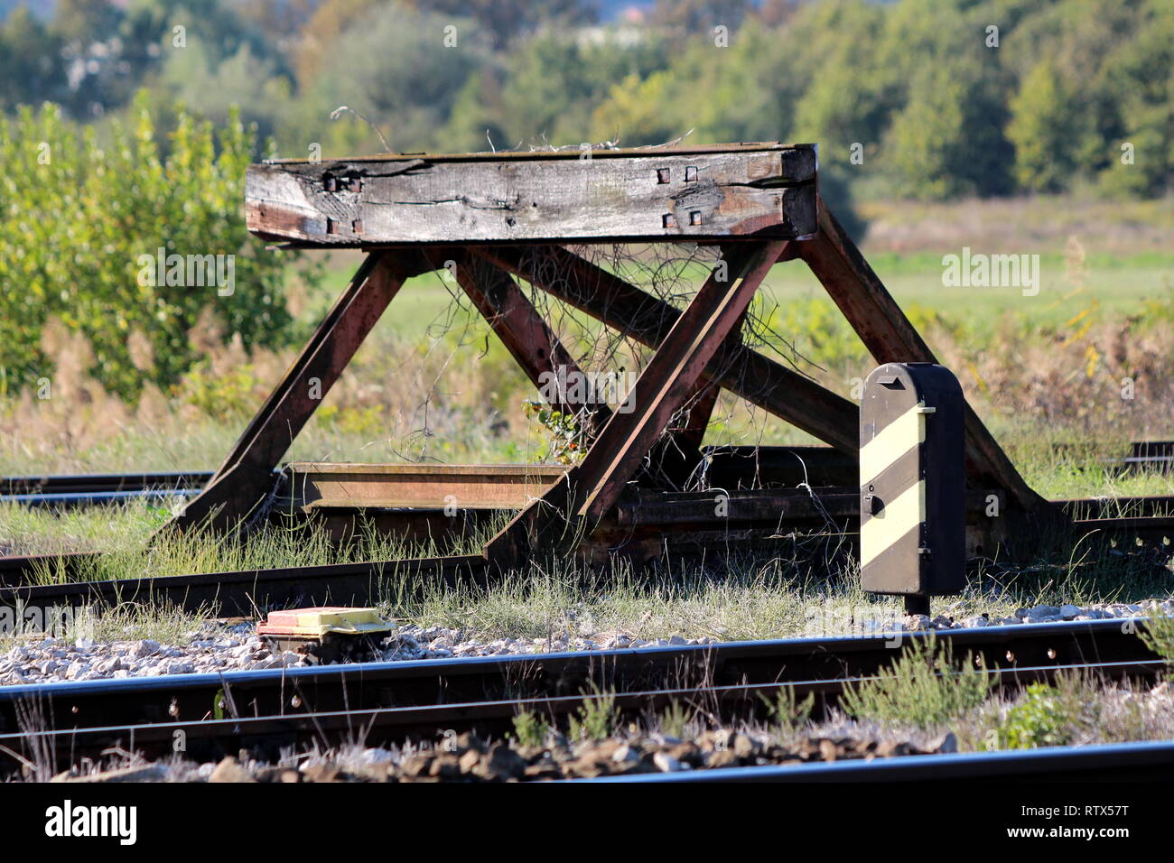 Railroad tracks stop barrier buffer at end of tracks in local train station with cracked wooden beam and rusted metal parts overgrown with dry plant Stock Photo