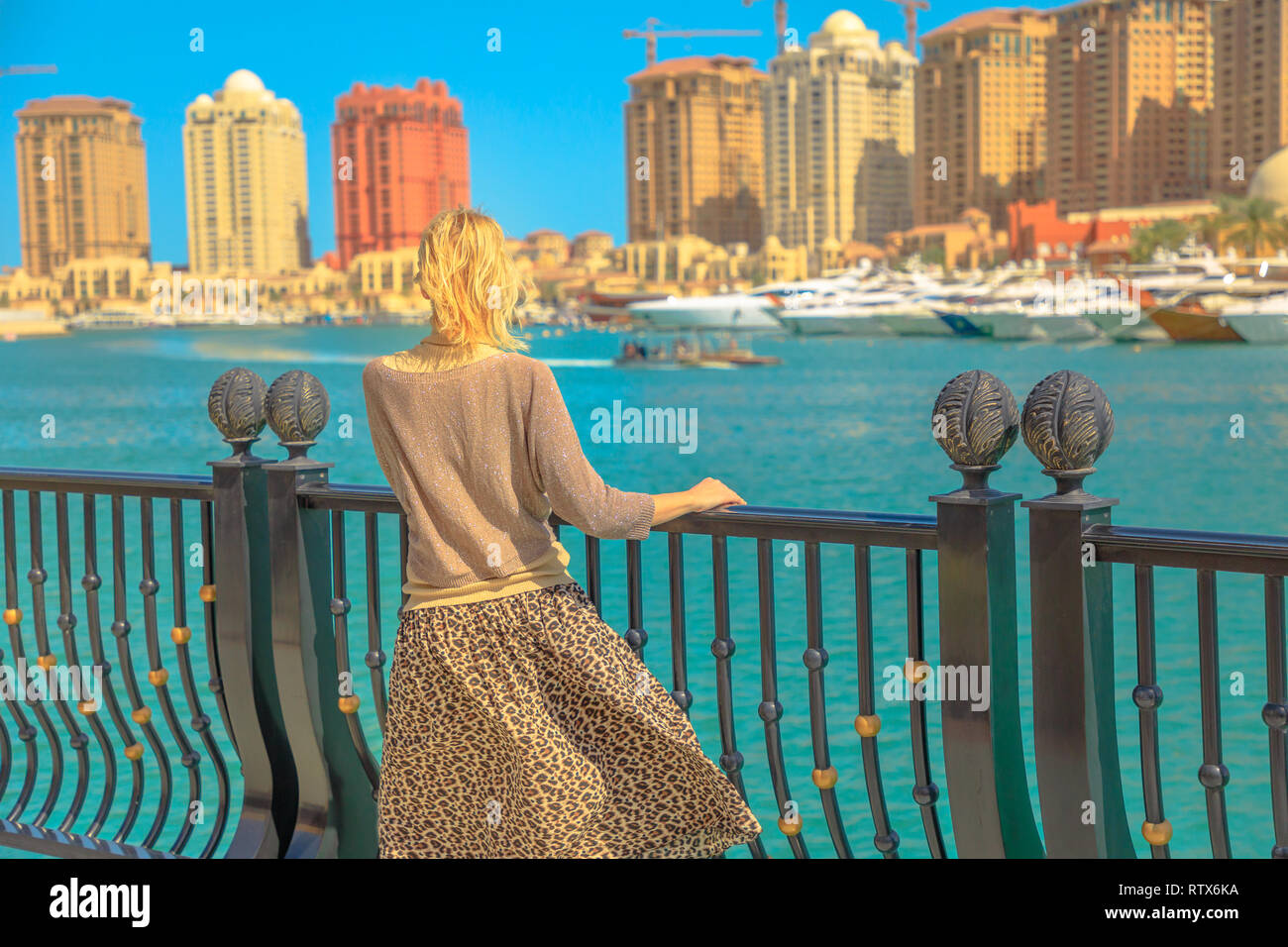 Tourism in Qatar. Blonde elegant woman at Marina walkway looks at Porto Arabia, The Pearl-Qatar's main harbor. Caucasian tourist traveler in Doha Stock Photo