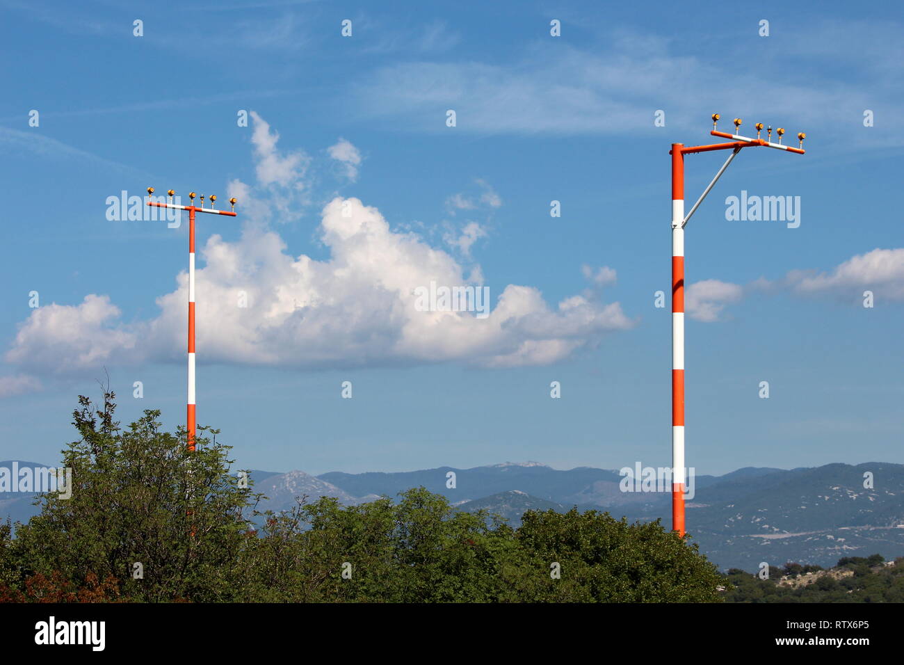 Two high metal red and white poles with multiple airport runway guiding lights rising high above dense trees with mountains and cloudy blue sky Stock Photo