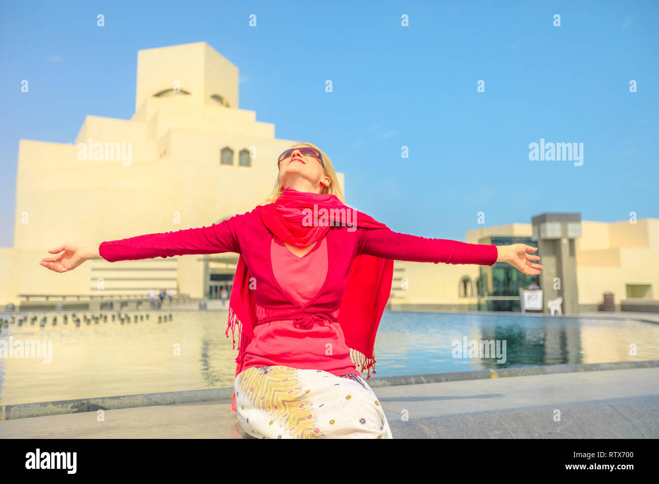 Carefree woman sitting in front of fountain of famous museum along the Corniche near Dhow Harbor in Qatari capital. Caucasian tourist enjoying in Doha Stock Photo