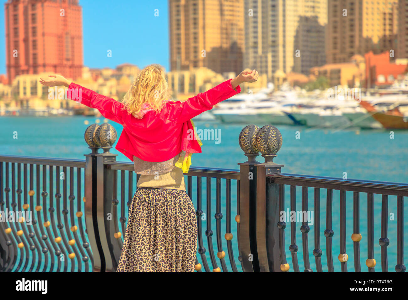 Tourism in Qatar. Blonde elegant woman at Marina walkway looks at Porto Arabia, The Pearl-Qatar's main harbor. Carefree tourist with open arms in Doha Stock Photo
