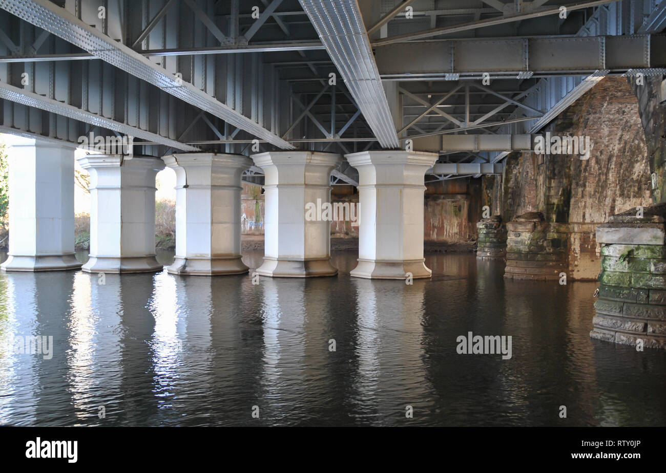 A view looking underneath a major railway bridge showing heavy steel column supports and their reflection in the water Stock Photo