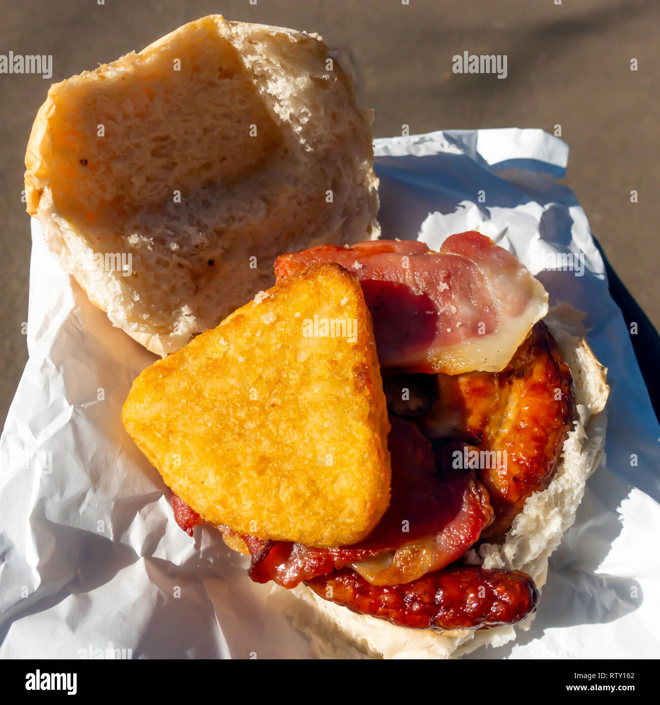 Lunchtime picnic snack a grilled Cumberland sausage sandwich with bacon and  a hash brown in white bread bun Stock Photo - Alamy