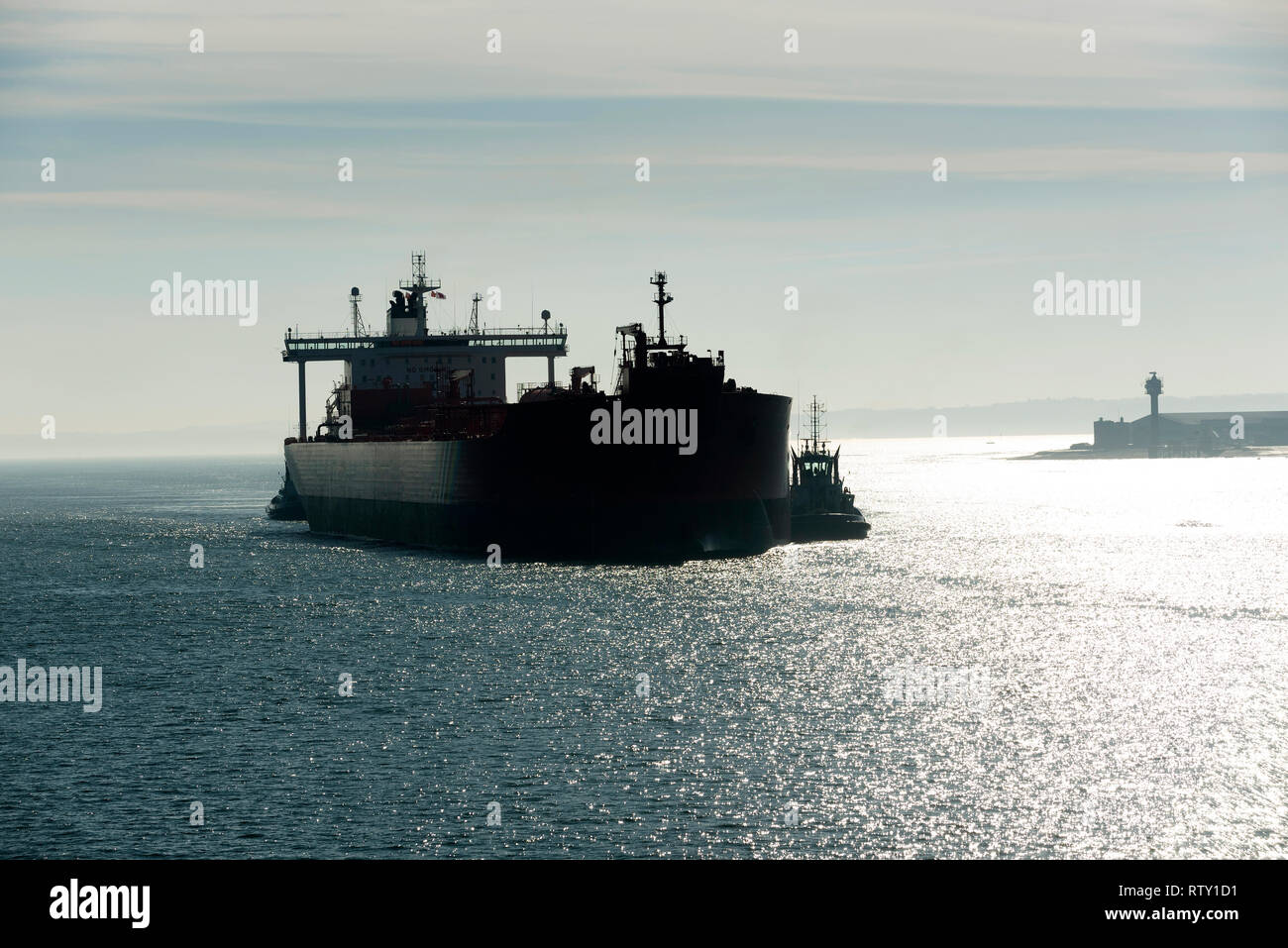 Southampton Water, Hampshire, England, UK. February 2019. Early morning against the light shot of a tug in attendance moving a broad beam oil tanker o Stock Photo
