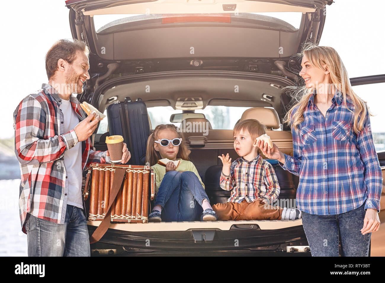 Mother, father, girl and boy loaded bags in the trunk of car. Parents stand near the car, having a lunch, family isready to travel. Stock Photo