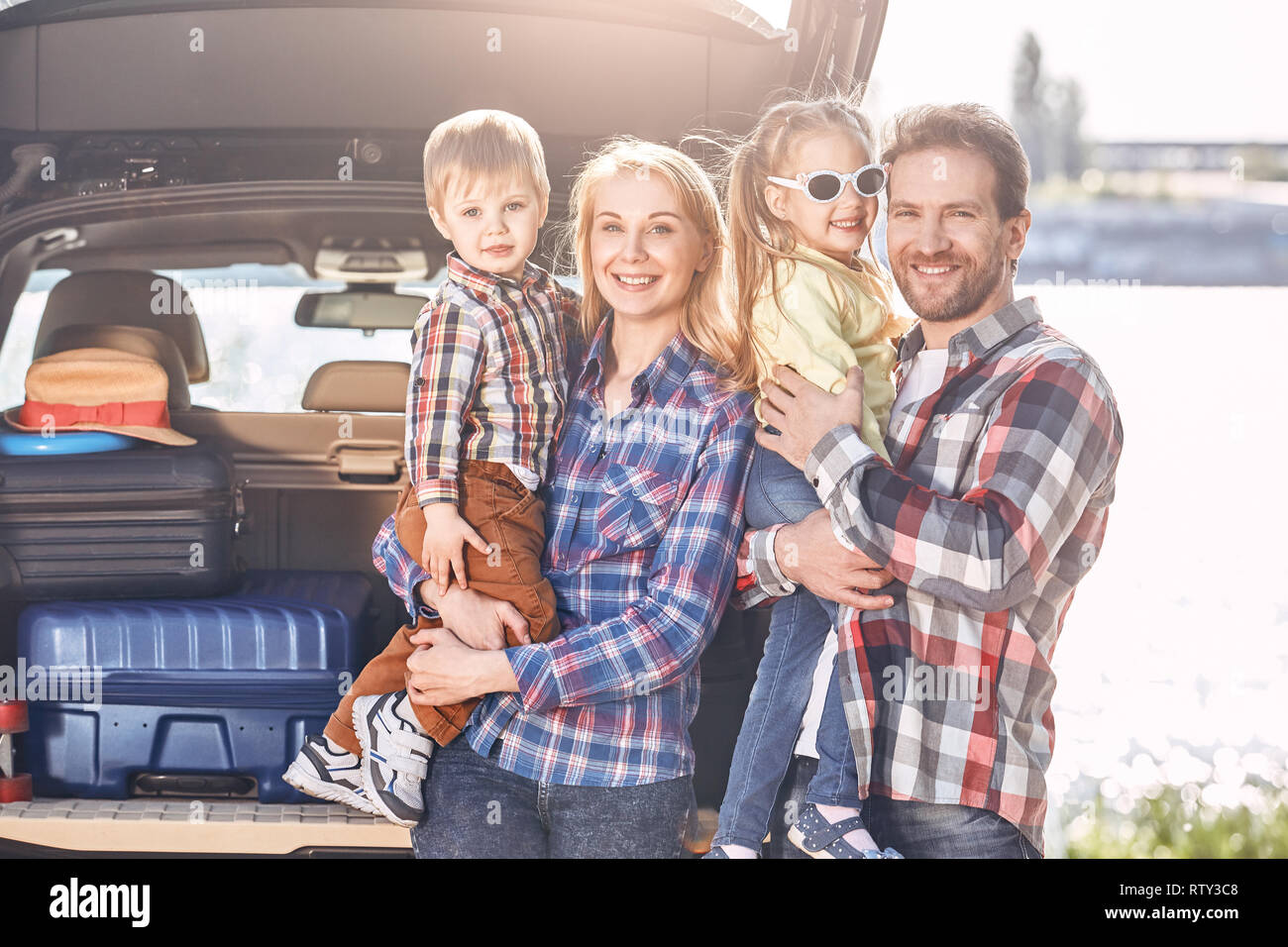 Mother, father, girl and boy loaded bags in the trunk of car. Parents stand near the car, holding their kids, they are ready to travel. Stock Photo