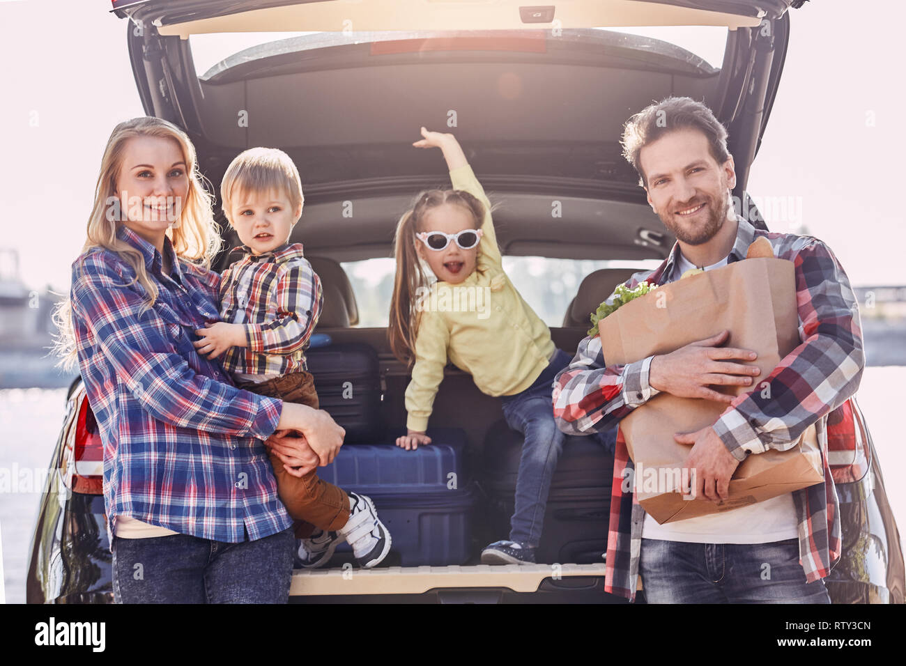 Mother, father, girl and boy are loading bags in the trunk of car. Family stand near the car, ready to travel. Stock Photo