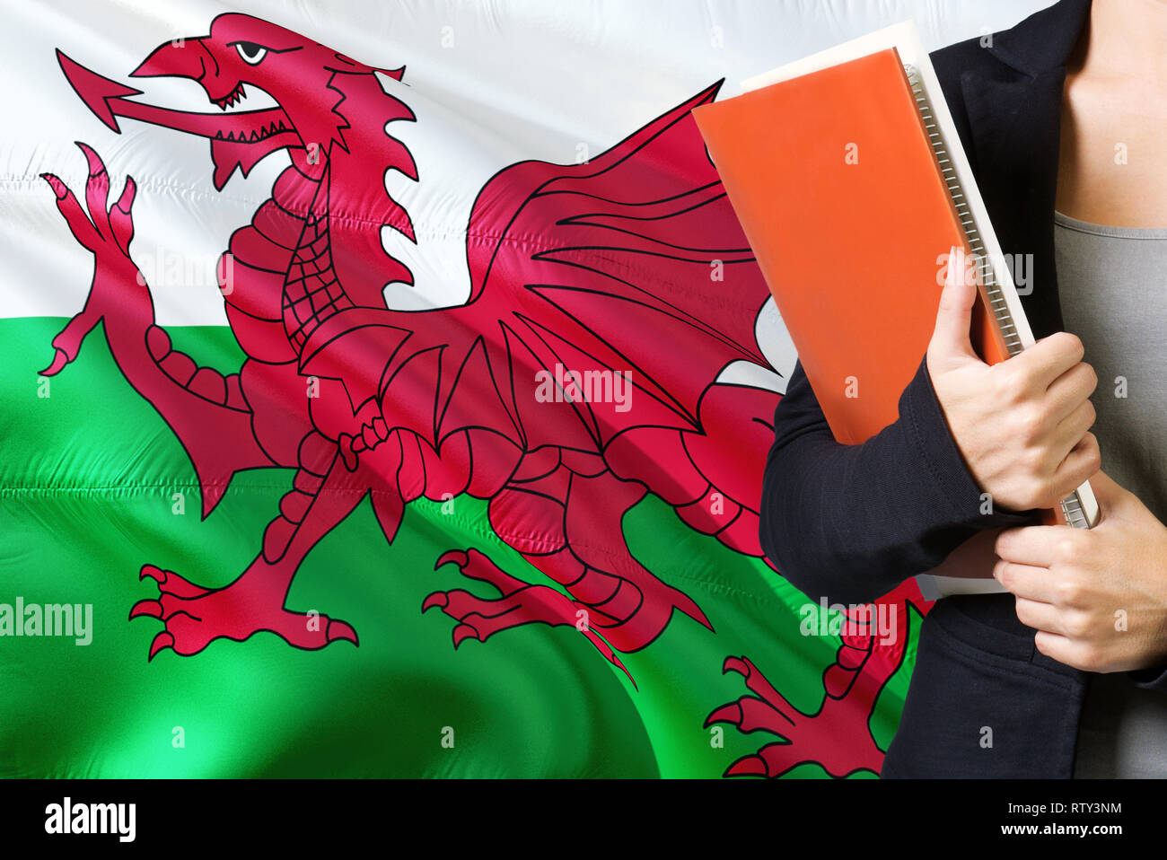 Learning Welsh language concept. Young woman standing with the Wales flag in the background. Teacher holding books, orange blank book cover. Stock Photo
