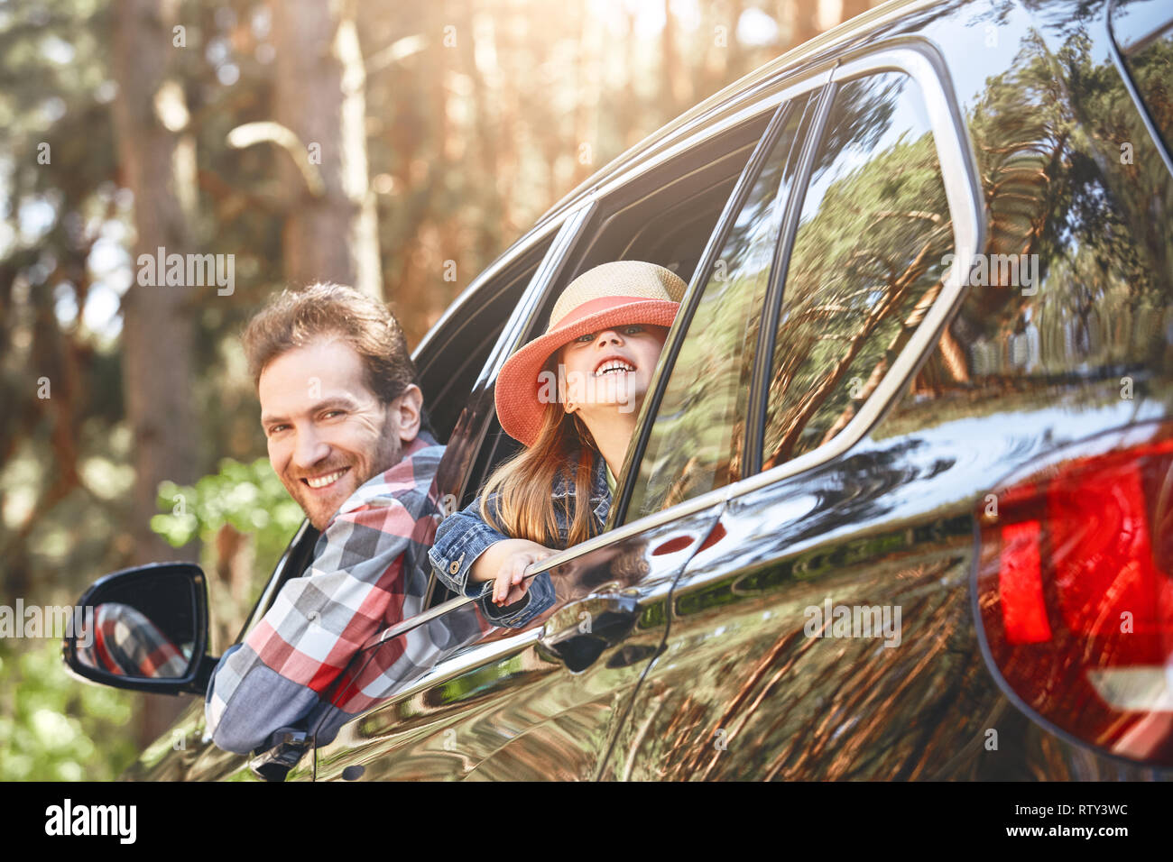 Cute little caucasian girl wearing a hat and her father in checkered shirt looking out the window while sitting inside the black shiny car. Man drivin Stock Photo