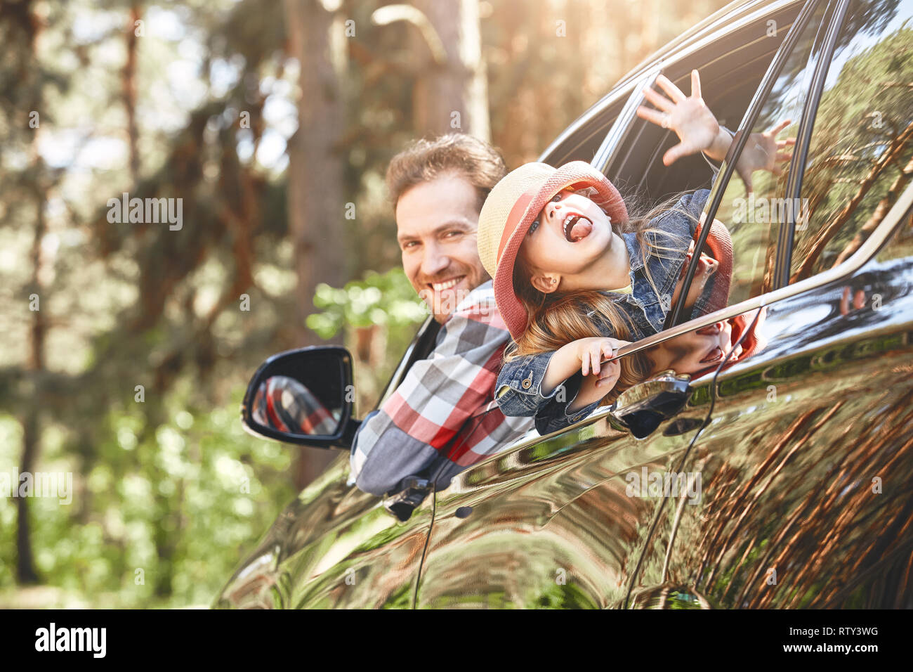 Cute little caucasian girl wearing a hat and her father in checkered shirt looking out the window while sitting inside the black shiny car. Man drivin Stock Photo