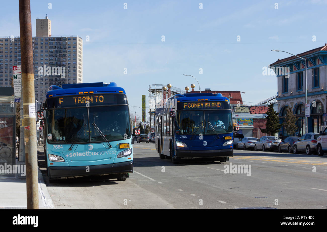 Like two halves of a couple, these Metropolitan Transportation Authority buses on Surf Avenue in Coney Island, Brooklyn, New York, appear to complete each other's thoughts, their digital/LED destination signs/indicators synching perfectly as the one in motion approaches the one that's parked, reading, from left to right, 'F TRAIN TO' and 'F CONEY ISLAND'. Structures appearing in the background of this peninsular community on the sunny winter February afternoon in 2019 include Brightwater Towers Condominium; Luna Park's historic wooden Cyclone roller coaster; and the Coney Island USA museum. Stock Photo