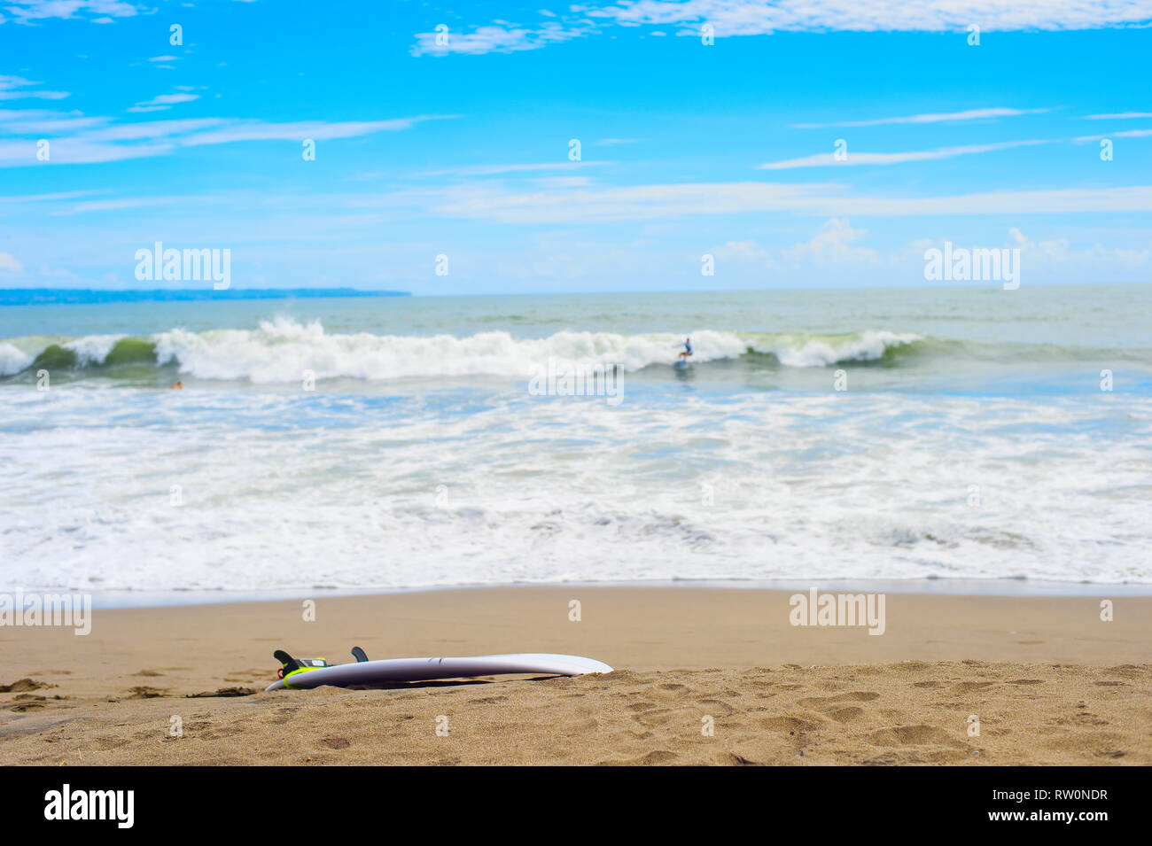 Surfboard on sandy beach, surfer riding on waves in background, Bali, Indonesia Stock Photo
