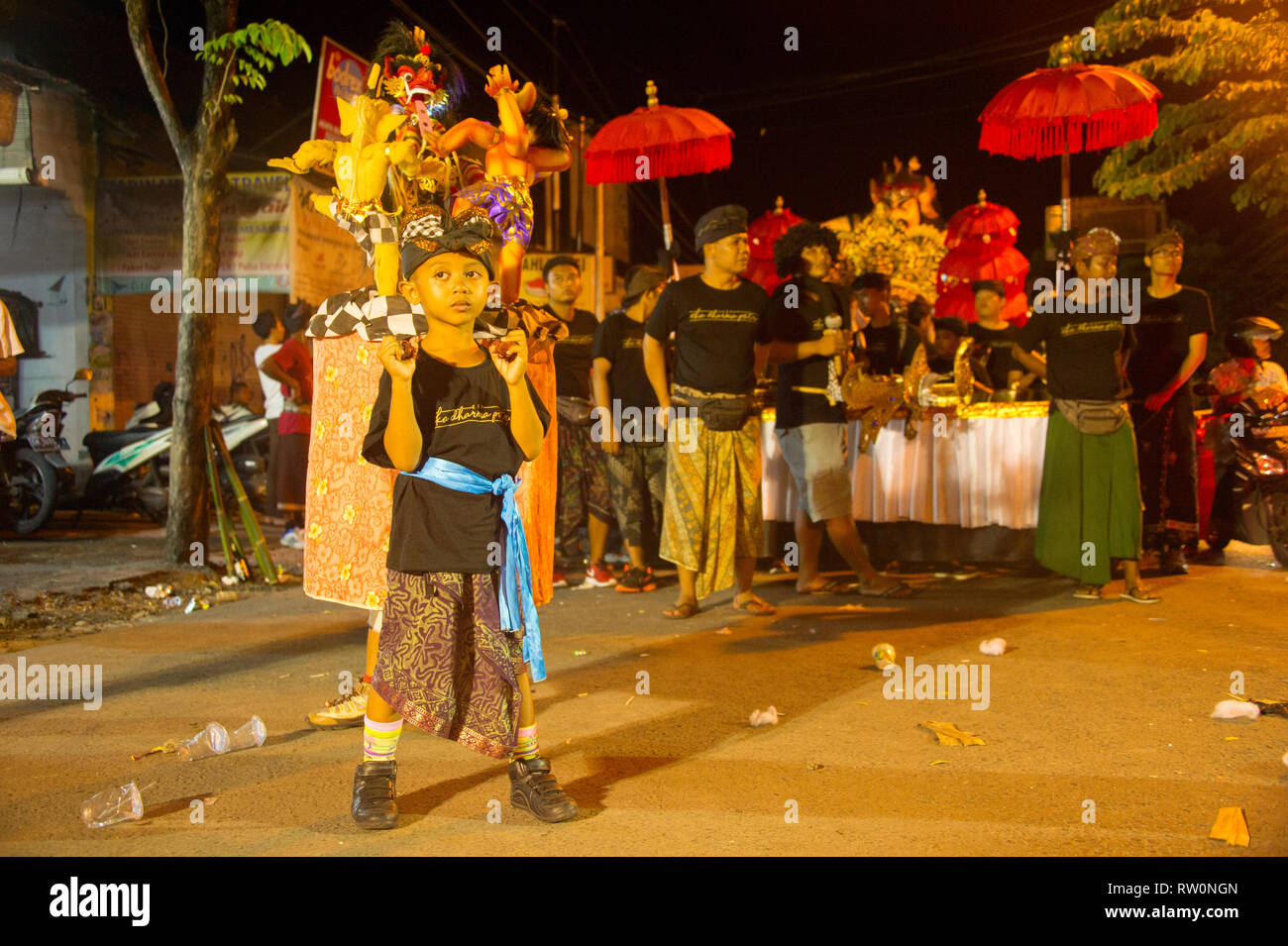 BALI, INDONESIA - MARCH 27, 2017: Boy in traditional costume holding Nyepi figures, traditional parade in evening Kuta street in background Stock Photo
