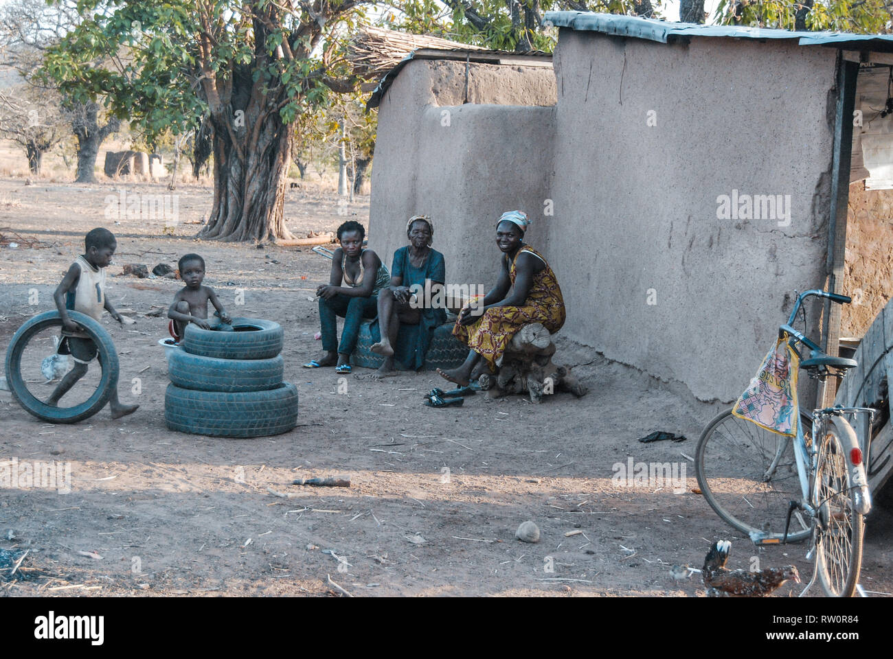 Three local Ghanaian women wearing traditional clothes are sitting in the yard and watching as their children play with some old car tyres. Stock Photo