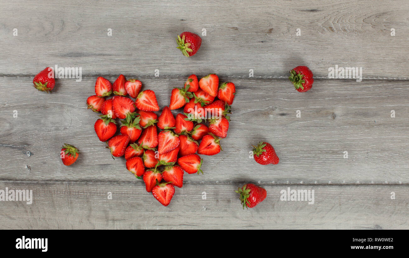 Strawberries cut to halves pieces, arranged in heart shape, on gray wood desk. Top down view. Stock Photo