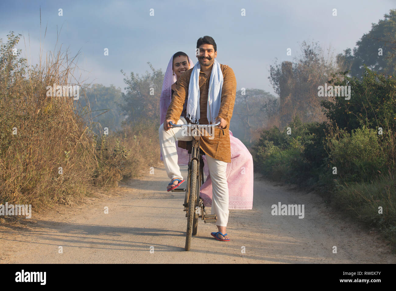 Happy rural couple in traditional dress riding on bicycle on a narrow ...