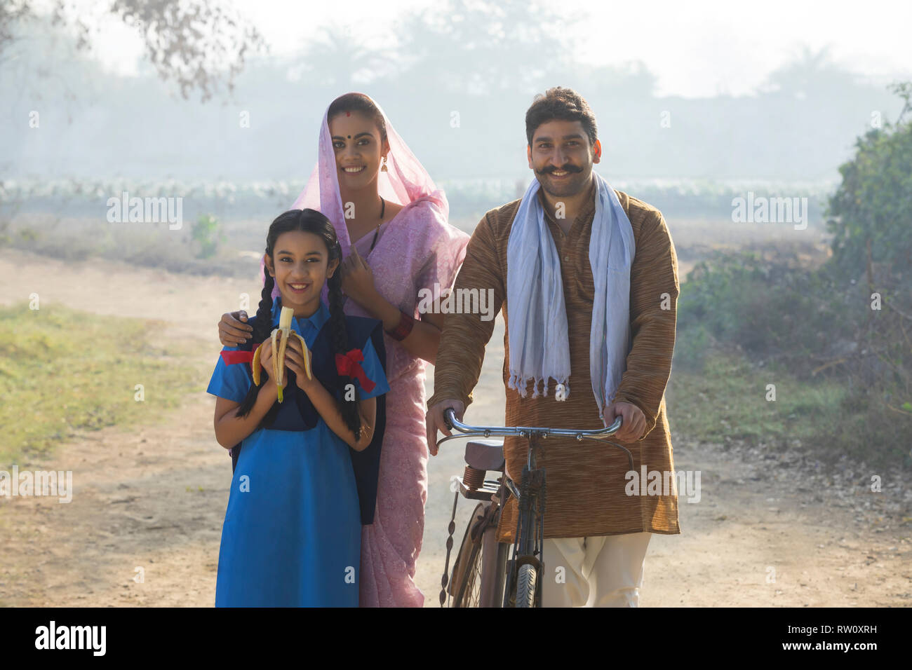 Happy rural family consisting of man, woman and a school going girl walking on pathway in a village. Stock Photo