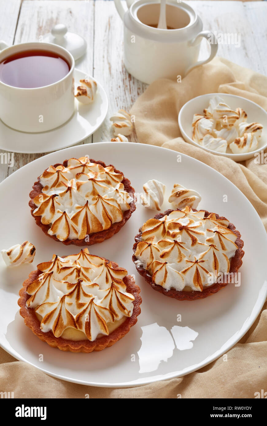 close-up of Lemon meringue mini pies with browned meringue peaks served with tea, mini marshmallows and brown organic sugar on wooden table with light Stock Photo