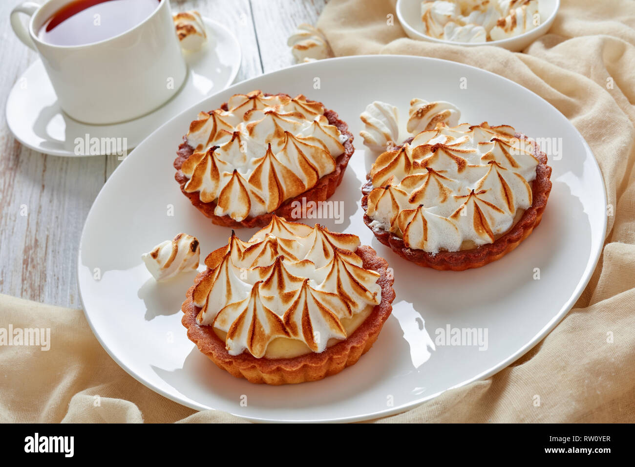 close-up of meringue mini tarts with browned meringue peaks served with tea, mini marshmallows and brown organic sugar on wooden table with light beig Stock Photo