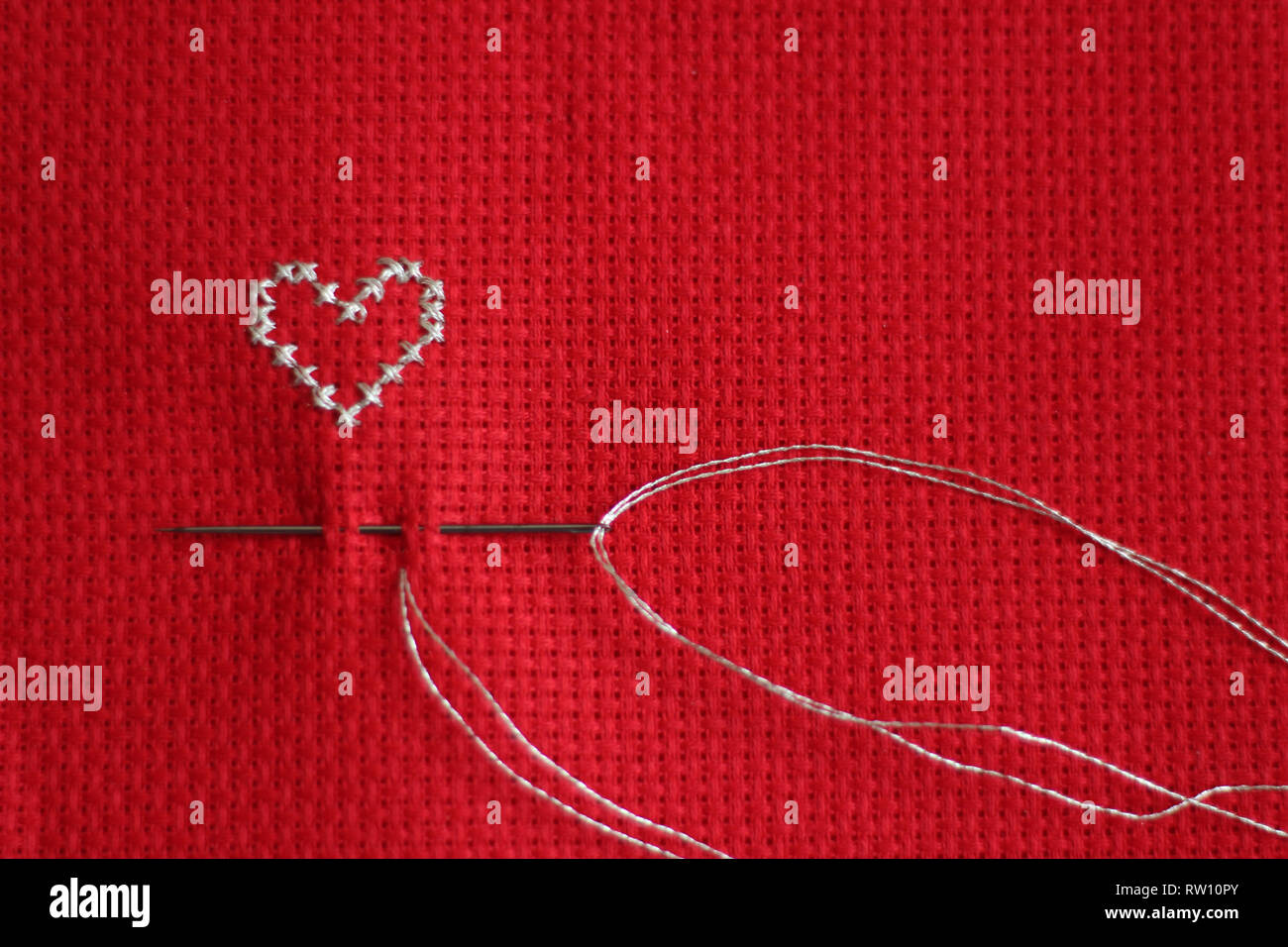 Silver cross stitched heart, needle and thread, red background Stock Photo
