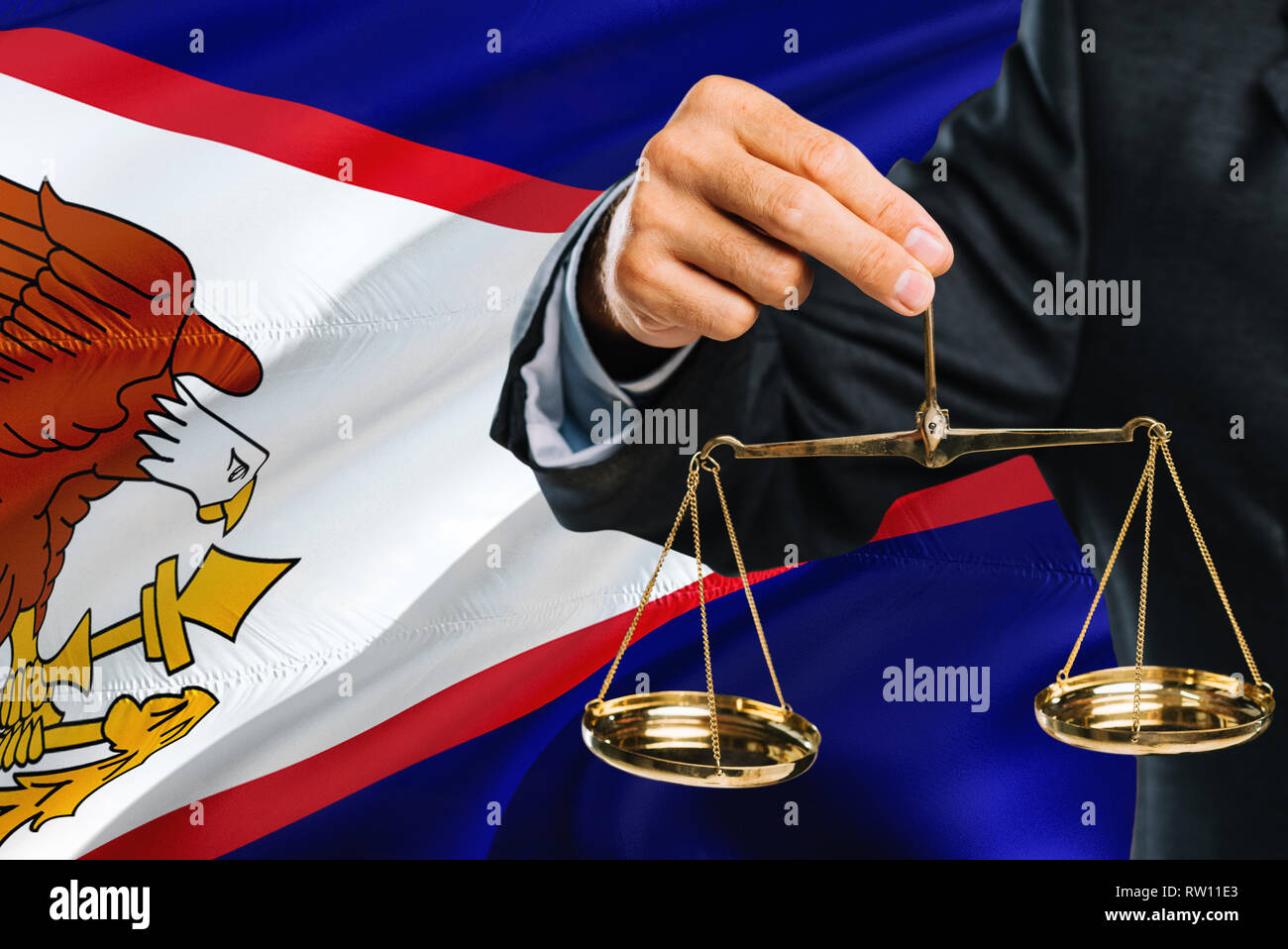 Judge is holding golden scales of justice with American Samoa waving flag background. Equality theme and legal concept. Stock Photo