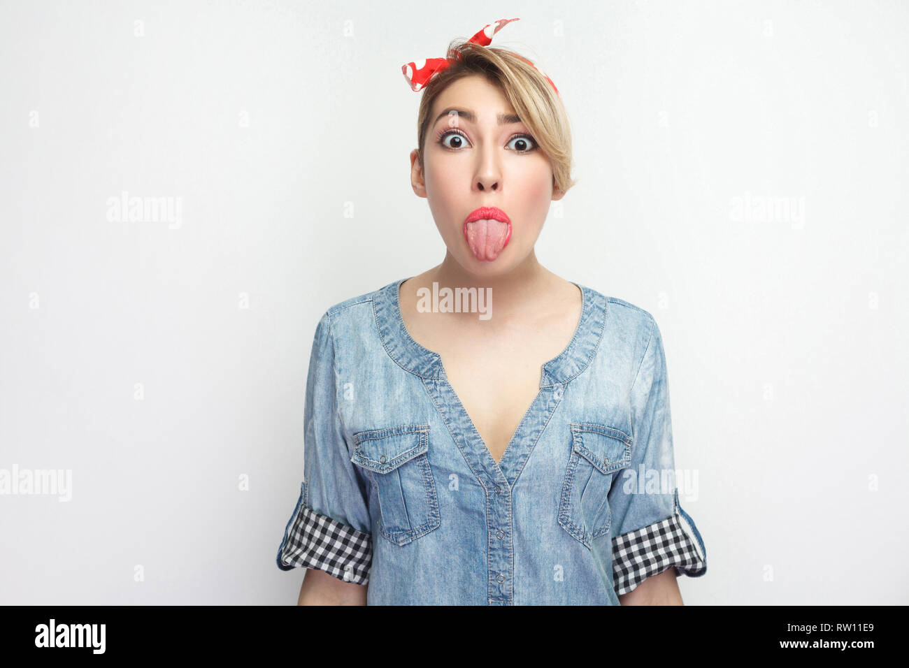 Portrait of funny beautiful young woman in casual blue denim shirt with makeup and red headband standing and looking at camera with tongue out and big Stock Photo