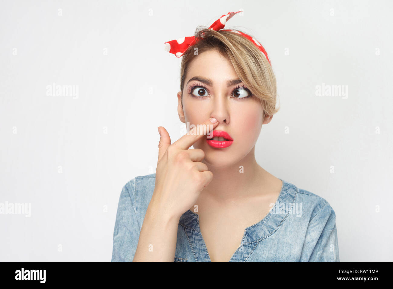 Closeup portrait of funny crazy crossed eyes young woman in casual blue denim shirt with makeup and red headband standing looking and picking her nose Stock Photo