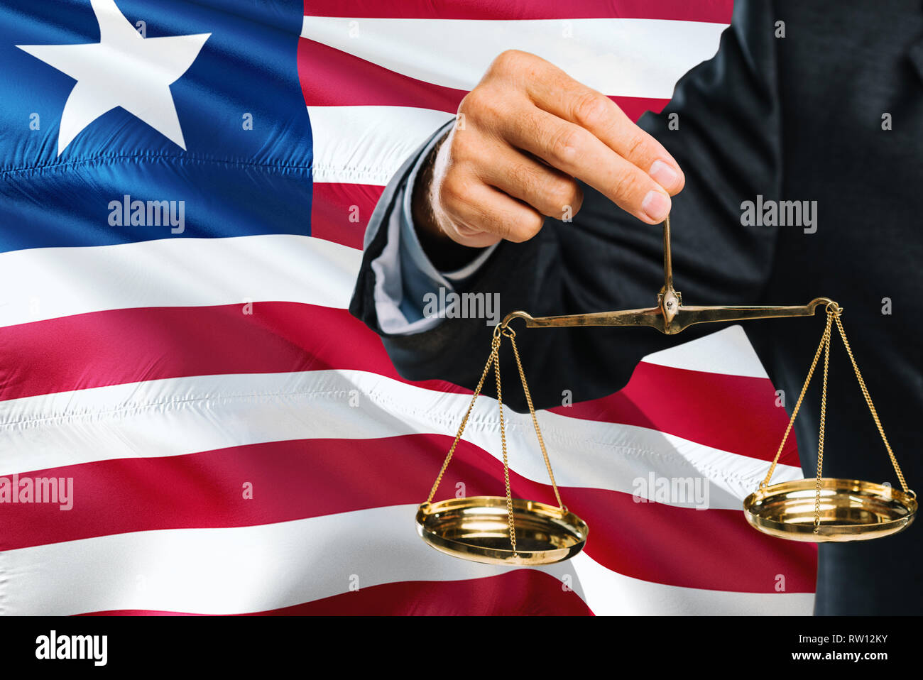 Liberian Judge is holding golden scales of justice with Liberia waving flag background. Equality theme and legal concept. Stock Photo